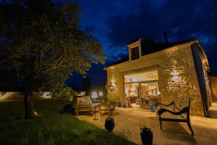 Casa de piedra iluminada por la noche con terraza acondicionada, mobiliario de exterior y grandes ventanales en Dordoña