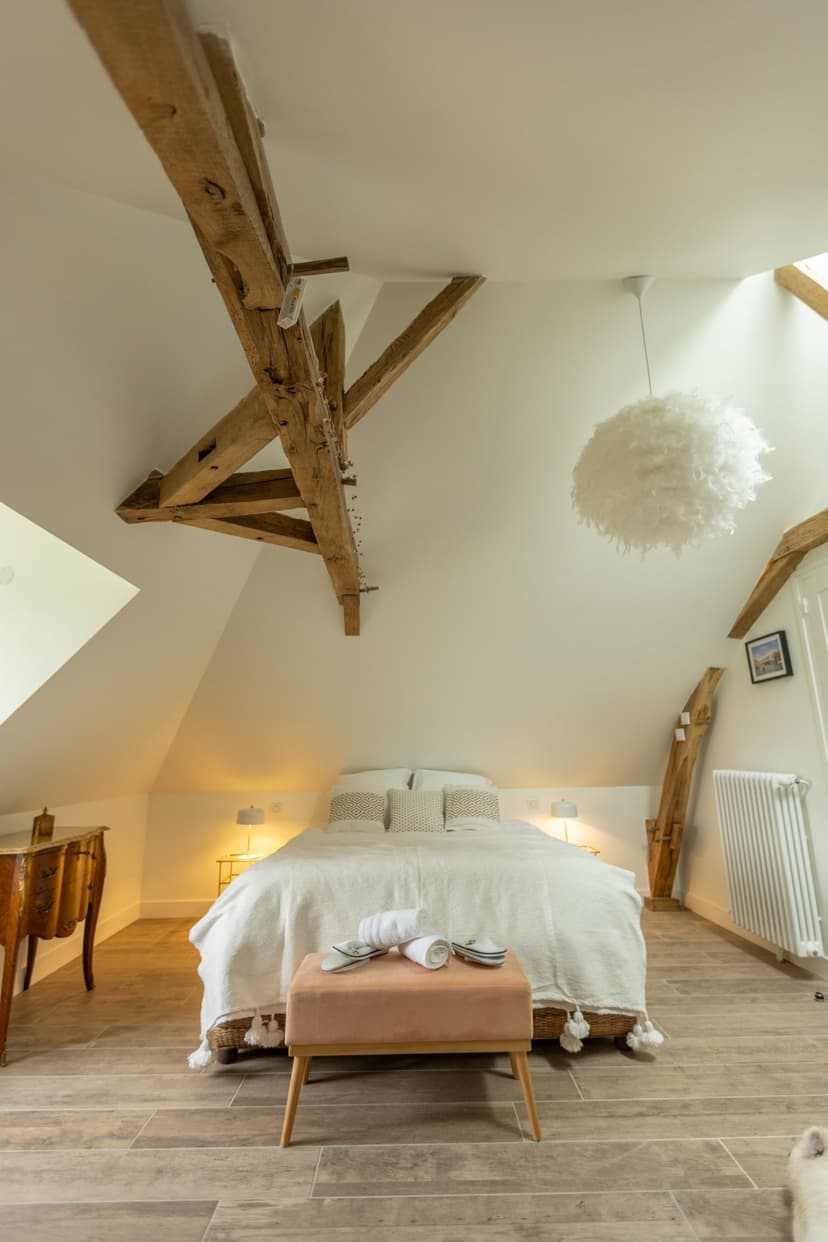Chambre élégante sous combles avec poutres en bois, lit double, suspension design et sol carrelage dans maison de vacances