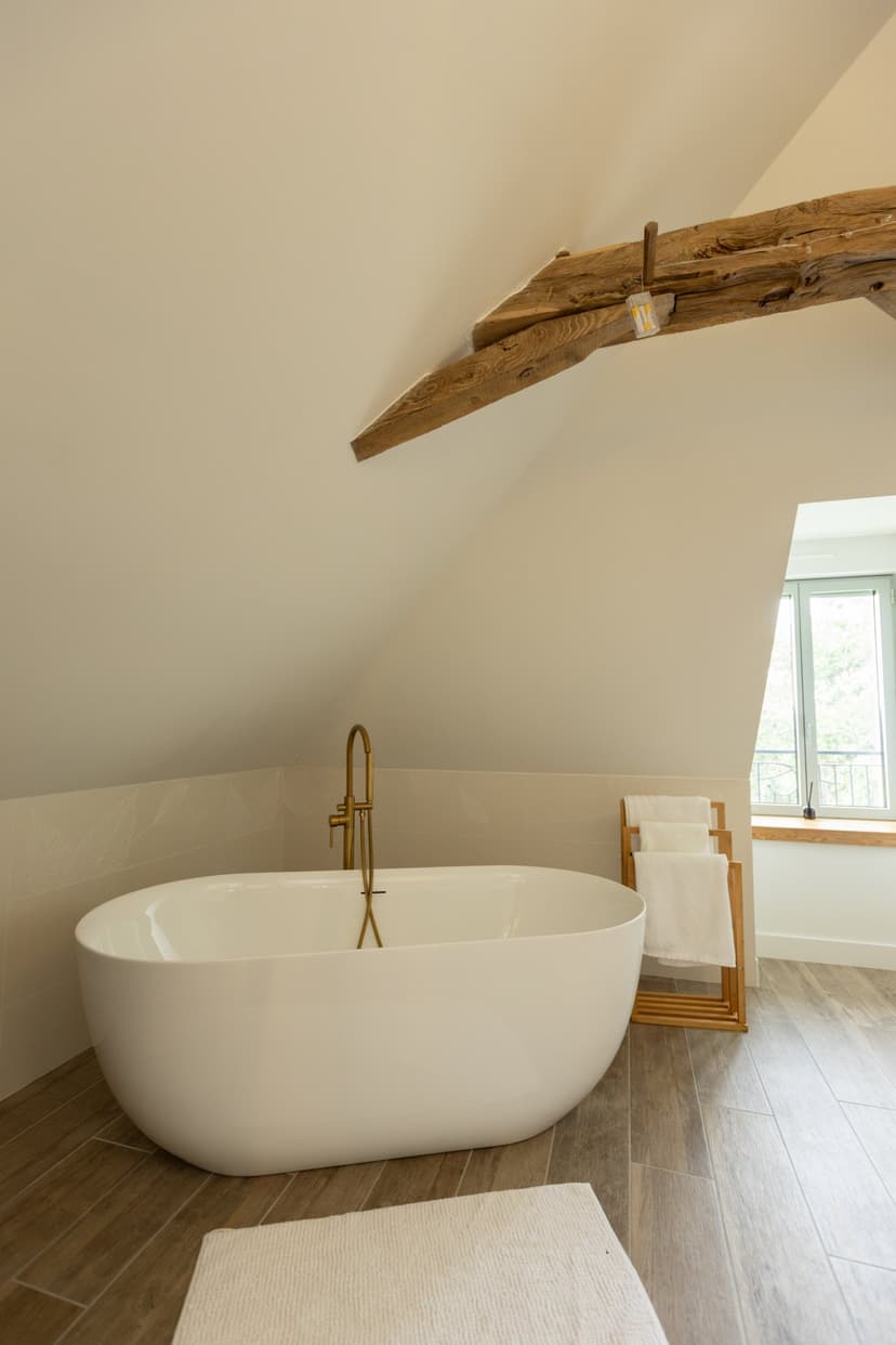 Modern white bathtub under wooden beam in attic bathroom with parquet flooring and gold fittings