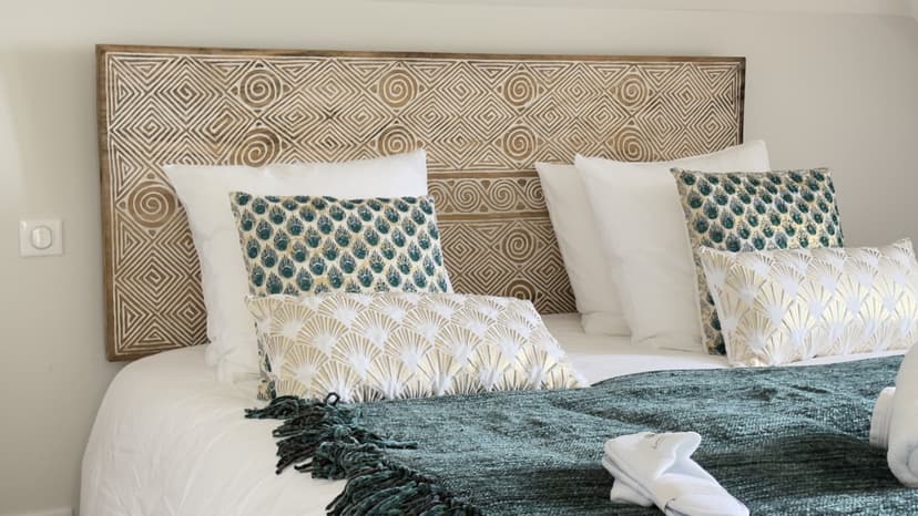 Chambre avec tête de lit en bois sculpté motifs géométriques, coussins verts et blancs, plaid vert sur lit blanc