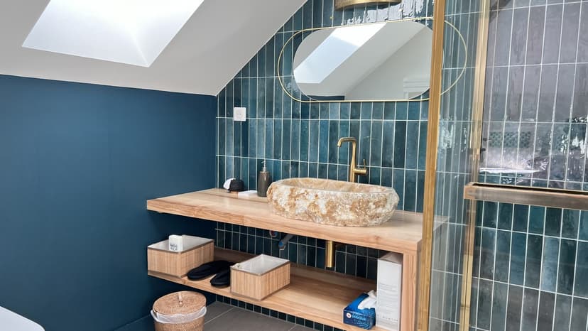Modern bathroom with blue-green tiling, stone basin, round gold mirror and wooden shelves under skylight