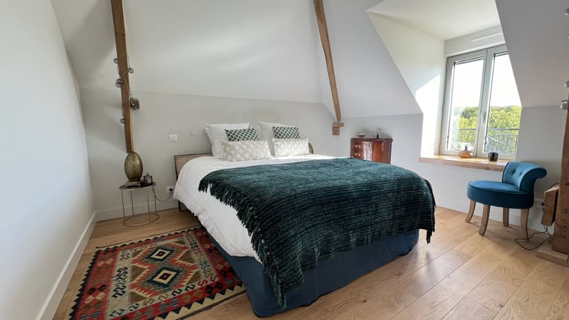 Chambre mansardée élégante avec lit double, tapis kilim coloré, fauteuil bleu et grande fenêtre avec vue campagne