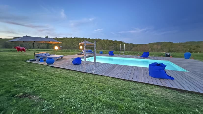Piscina iluminada al atardecer con deck de madera, mobiliario azul y sombrilla en propiedad vacacional de Dordoña