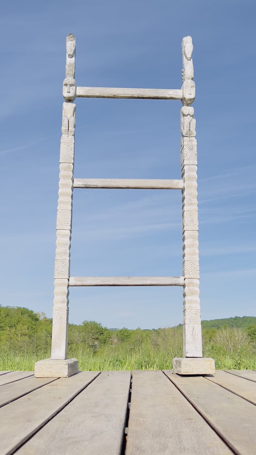 Gran escultura de madera representando una escalera con figuras talladas en terraza con vistas al campo