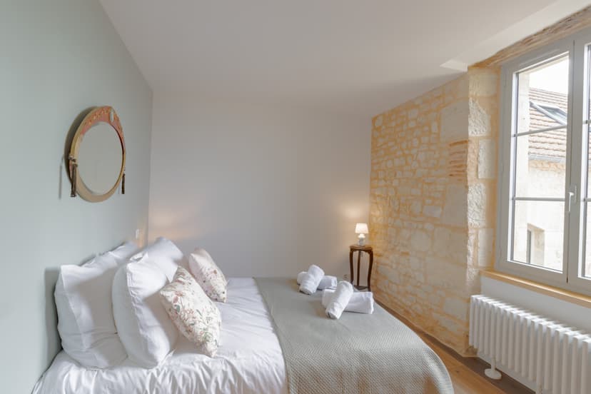 Elegante slaapkamer met wit bed, natuurstenen muur, ronde spiegel en groot raam met uitzicht op pannendaken