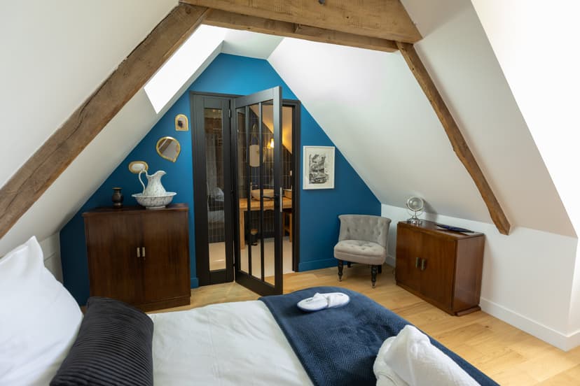 Quarto elegante sob mansarda com vigas de madeira, parede azul, banheiro envidraçado e mobiliário vintage na Dordonha